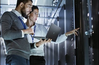 A man holding a laptop standing next to a woman pointing at a control panel