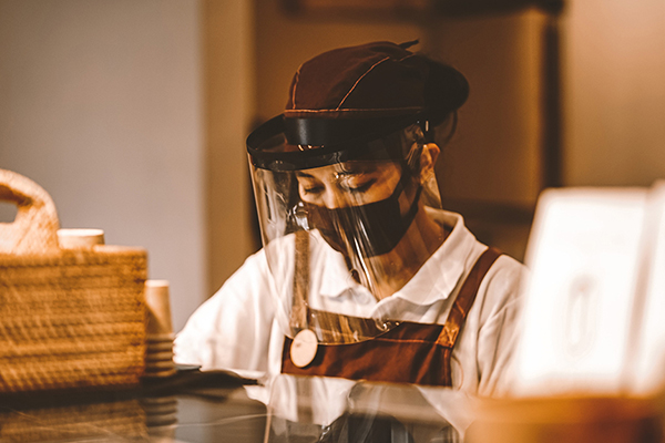 A barista wearing a face-mask and face-shield, working behind a counter